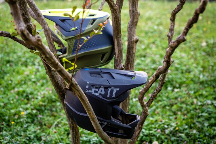 Two helmet models from Leatt hanging on a tree branch, surrounded by a grassy area. The top helmet has a vibrant design in green and gray, while the bottom helmet is sleek and black with a logo.