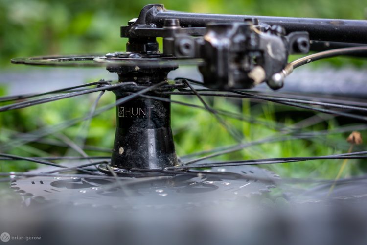 Close-up image of a bicycle wheel hub with spokes, featuring a black component labeled "HUNT." The background is blurred greenery, suggesting an outdoor setting.