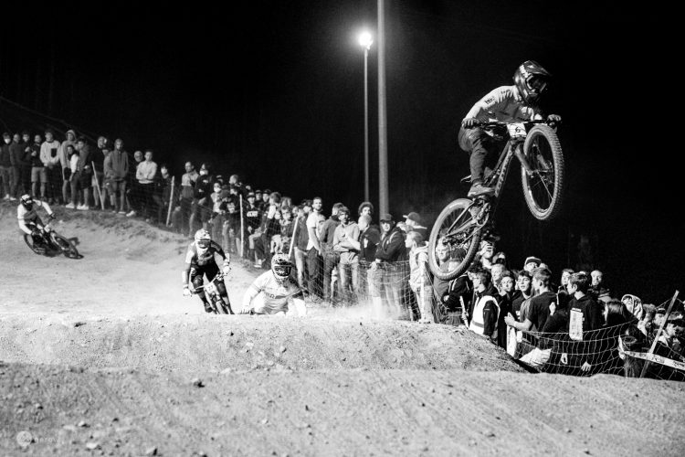 A black and white image of a night-time mountain biking event, showcasing riders in action on a dirt track. One cyclist is performing a jump over a ramp, while two others are racing on the track below. A crowd of spectators watches from the sidelines, captivated by the thrilling competition. Dust is kicked up from the track, adding to the dynamic atmosphere of the event.