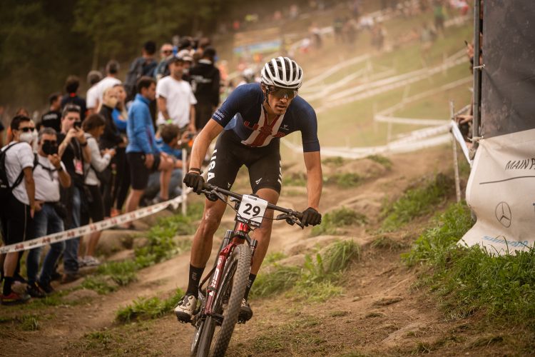 A male mountain biker wearing a blue jersey with a U.S.A. emblem races along a dirt path, surrounded by spectators. Dust rises from the ground as he navigates a turn, showing focused determination. The background features a line of fans watching the event.