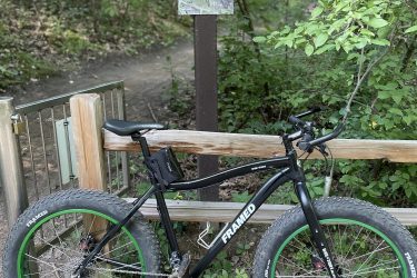A black mountain bike with green wheels is parked next to a wooden trail marker at Lone Lake Park, which features a map of the Multi-Use Mountain Bike Trail. The surrounding area is lush with greenery, suggesting a scenic outdoor setting. Lone Lake Park mountain bike trail.