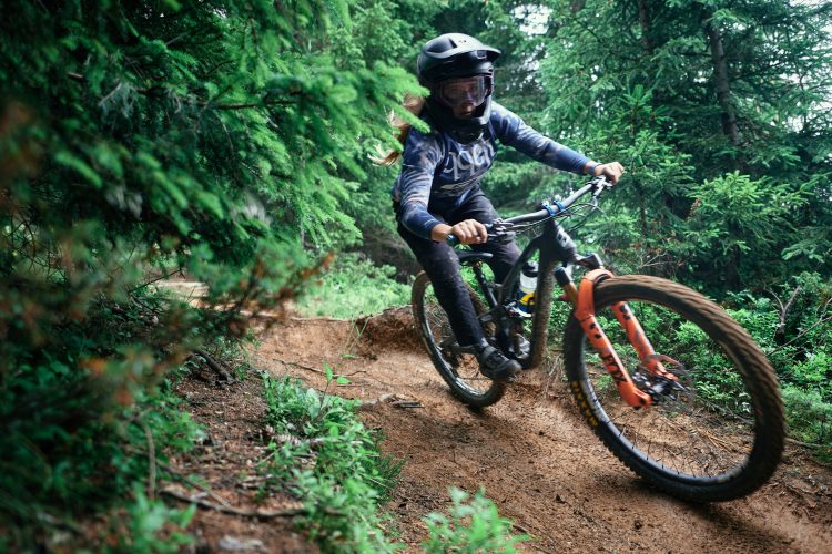 A mountain biker navigating a dirt trail through a lush forest. The rider, wearing a helmet and protective gear, leans into the turn as they accelerate on a rocky path surrounded by green trees and vegetation.