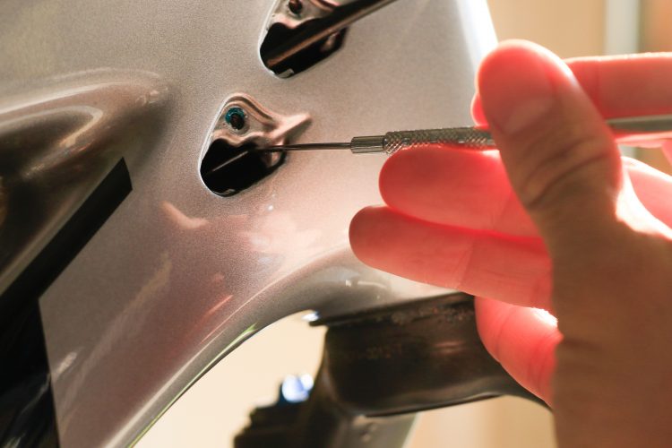 A close-up image of a person's hand holding a precision tool while working on a shiny metallic surface. The tool is inserted into a small opening on the surface, which appears to be part of a larger mechanical or automotive component. Soft lighting enhances the metallic finish and the details of the hand and tool.