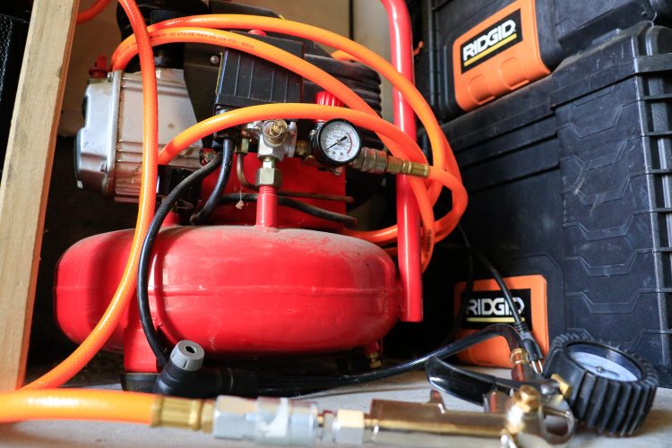 A red air compressor with orange hoses and gauges, surrounded by black storage cases with orange labels, situated in a workshop setting.