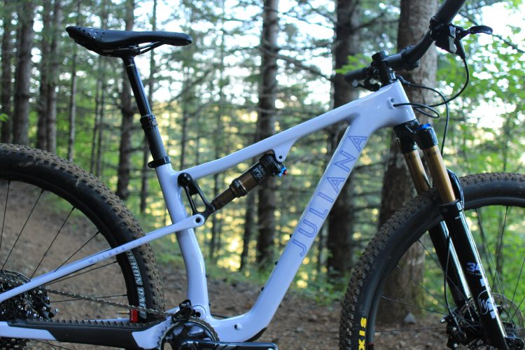 A close-up view of a white mountain bike with the word "Juliana" visible on the frame. The bike features a suspension system and is set against a backdrop of a forested trail with trees and greenery in the background.