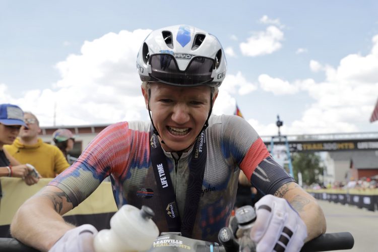 A tired mountain biker in a colorful cycling jersey and helmet is at the finish line, holding onto his bike and water bottle. He has a medal around his neck and is smiling while appearing exhausted. Spectators can be seen in the background, celebrating the race. The sky is partly cloudy, suggesting an active outdoor event.