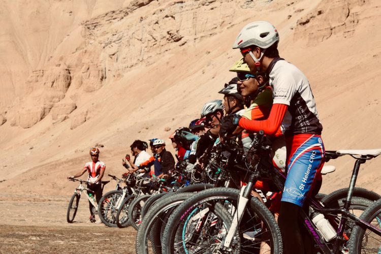 A group of mountain bikers lined up on their bikes, wearing helmets and cycling gear, with a barren landscape of sandy hills in the background. The riders are focused and appear ready for a race or a cycling event.