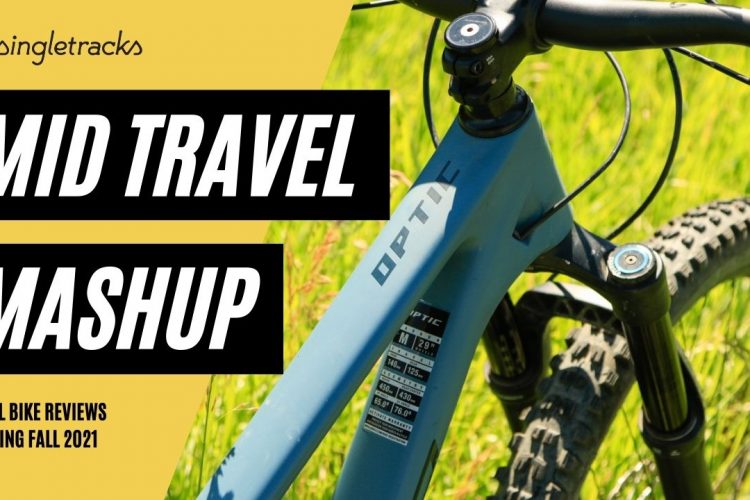 Alt text: Close-up of a blue mountain bike frame with "Optic" branding, featuring mid-ride components. The background shows green grass, and bold text overlays read "Mid Travel Mashup" and "Trail Bike Reviews Coming Fall 2021."