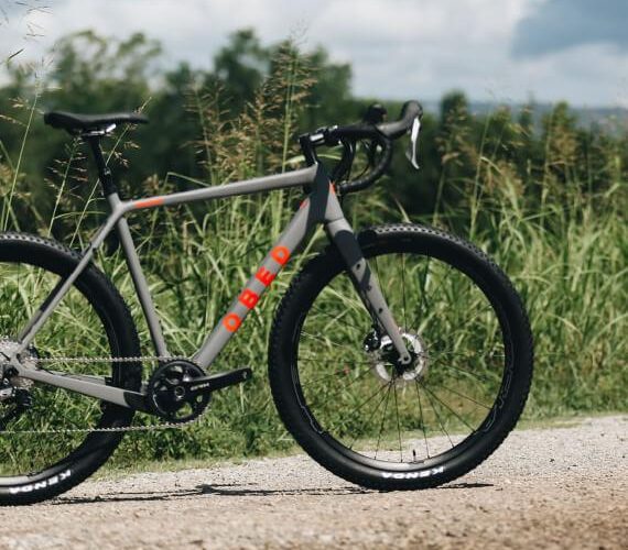 A gray bicycle with red accents is parked on a gravel path surrounded by tall grass and greenery under a partly cloudy sky. The bike features a modern design, with drop handlebars and thick tires suitable for off-road riding.