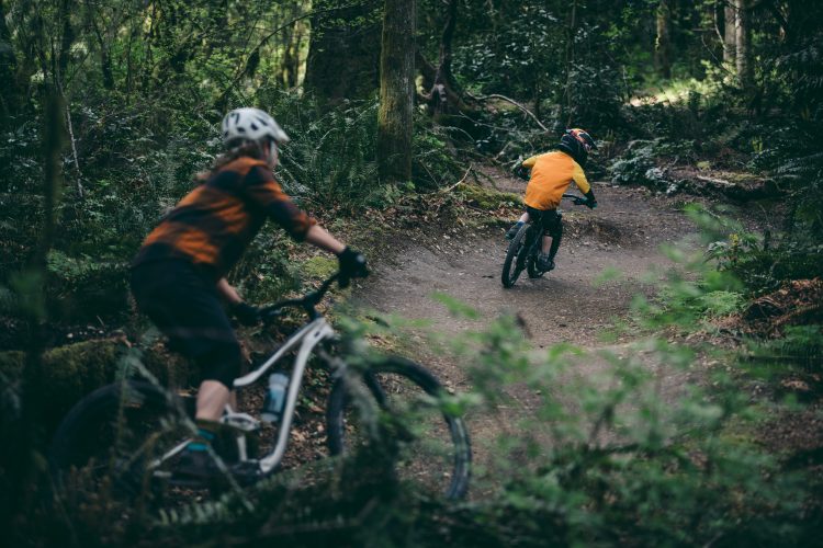 A child in an orange shirt rides a mountain bike along a dirt trail in a lush green forest. In the foreground, an adult cyclist in a striped shirt and helmet is positioned slightly to the left, observing the child as they navigate a bend in the trail. The scene captures the excitement of biking in nature surrounded by trees and foliage.