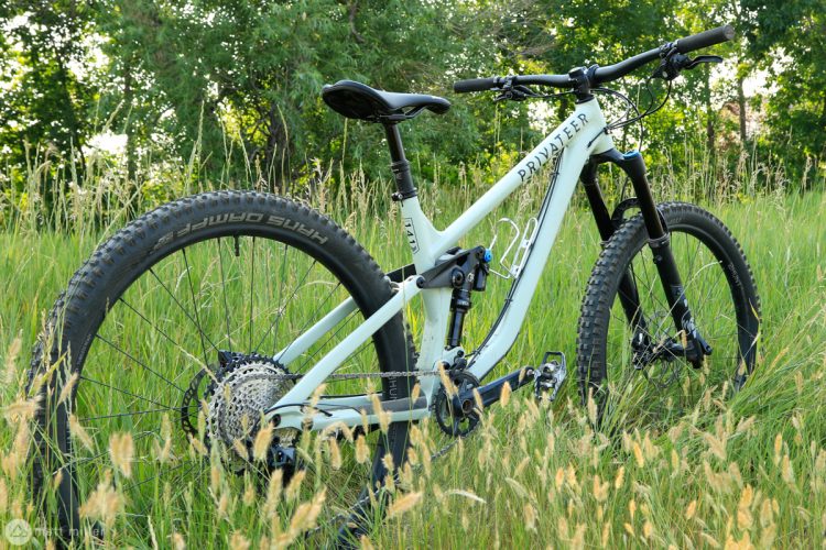 A light blue mountain bike resting amidst tall green grass and wildflowers, showcasing its sleek frame, wide tires, and suspension components. The backdrop features trees, creating a natural outdoor setting.