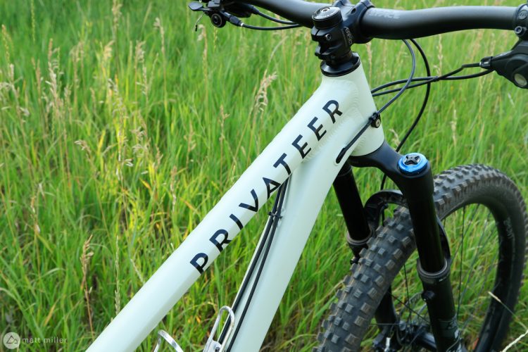 Close-up view of a light-colored mountain bike frame with the word "PRIVATEER" prominently displayed. The bike's front fork and handlebars are visible, and the background consists of lush green grass, creating a natural outdoor setting.