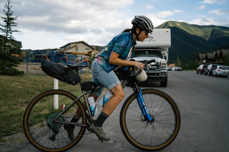 A cyclist with a multi-colored shirt and helmet is maneuvering a bicycle along a street surrounded by mountains, while carrying gear in bags attached to the bike. The background features parked vehicles and a mix of greenery and buildings under a partly cloudy sky.