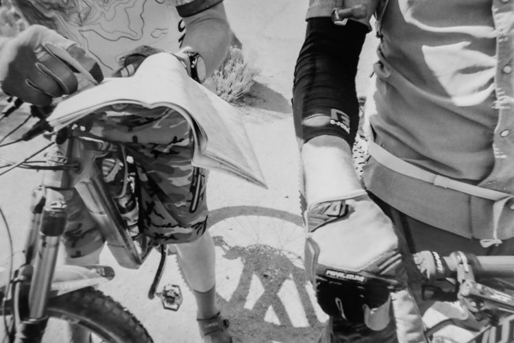 Two cyclists, one holding a map and the other adjusting their gear, gathered around a mountain bike. The scene captures their focus on navigation in an outdoor setting, with details including camouflage shorts and biking gloves visible. The background shows a faint shadow of a bicycle wheel on the ground.