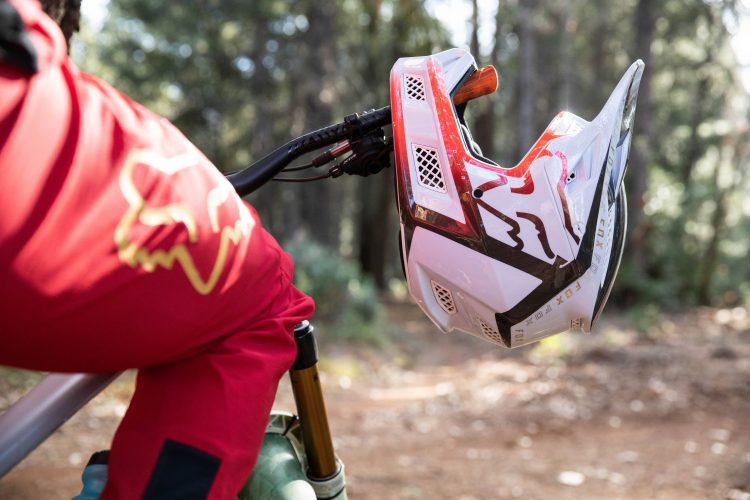 A close-up view of a mountain bike helmet positioned on the handlebars of a bike, with a rider wearing a red jacket in the background. The scene is set in a forest, featuring a trail with dirt and greenery.