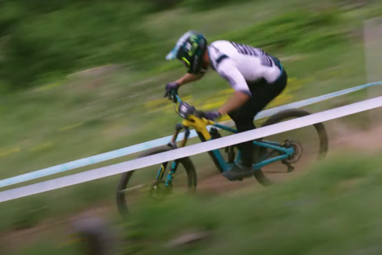 A mountain biker riding swiftly along a dirt trail, leaning into a turn with a blurred background, showcasing speed and action amidst a green landscape. The bike is teal with yellow accents, and the rider is wearing a helmet and protective gear. A white race tape marks the course boundary.