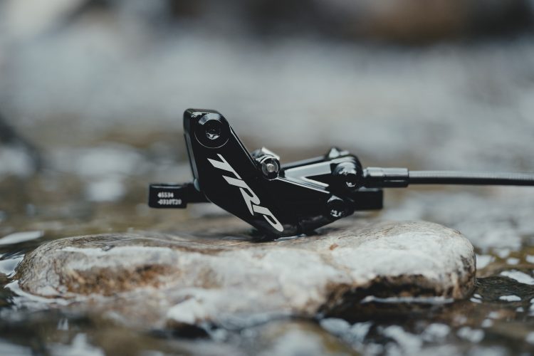A black TRP bicycle brake caliper resting on a smooth stone in shallow water, with a blurred natural background.