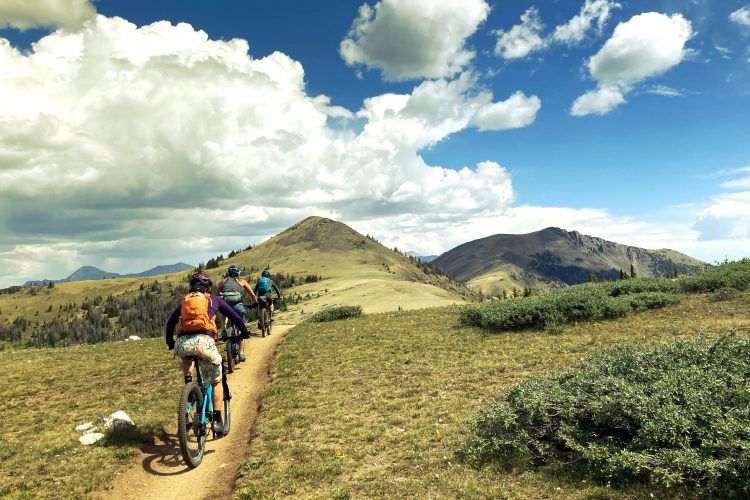 A group of mountain bikers riding along a dirt trail in a scenic landscape, surrounded by rolling hills and a blue sky with fluffy clouds. The trail winds through green grass and sparse shrubbery, leading towards a distant mountain range.