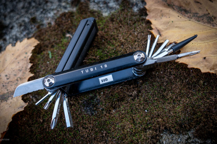 A multi-tool with various attachments, including a knife blade and screwdriver tips, resting on a bed of moss with a dried leaf nearby. The tool is marked with the label "TUBI 18."