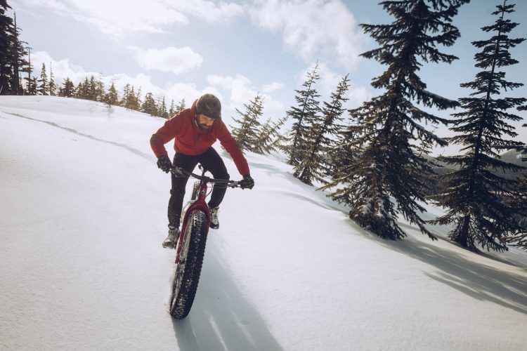 A person riding a fat bike on a snow-covered trail surrounded by evergreen trees, with a bright sky and scattered clouds in the background. The cyclist is wearing a red jacket and a helmet, focused on navigating the snowy terrain.