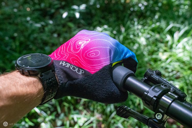 A close-up of a gloved hand gripping a bicycle handlebar, with a colorful glove featuring a pink and blue topographic design. A digital watch is visible on the wrist, along with parts of the bike's braking system, set against a background of green foliage.
