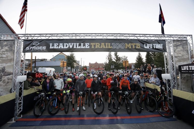 A large group of mountain bikers gathered at the starting line of the Leadville Trail 100 MTB race, under a banner displaying the event name. Participants, dressed in various cycling gear and helmets, are positioned with their bicycles, with an electronic timer showing the countdown. The backdrop includes trees and buildings, set against an early morning sky.