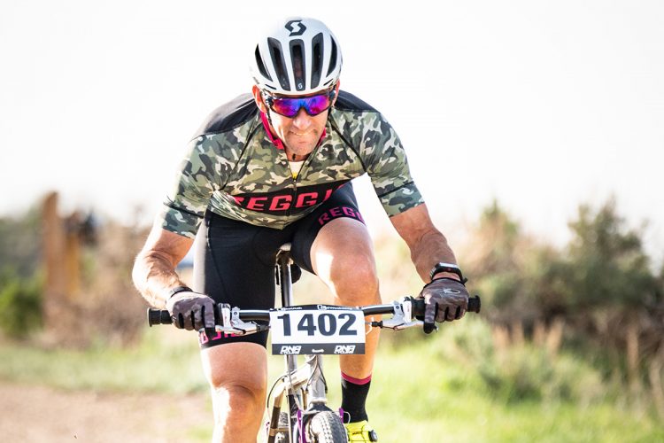 A male cyclist wearing a camouflaged jersey and sunglasses is intensely focused while racing on a mountain bike. The number "1402" is displayed prominently on his bike. He is in a grassy area with a blurred background suggesting a competitive outdoor setting.