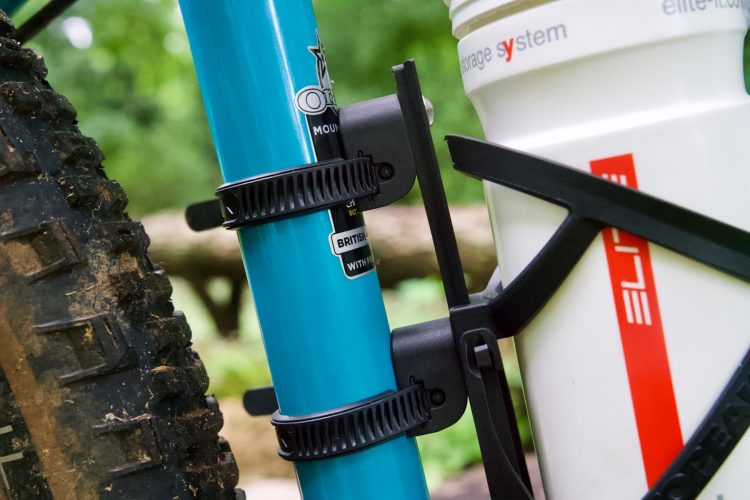 Close-up view of a turquoise bicycle frame with a water bottle mounted on the side. The image shows black straps securing the water bottle cage to the frame, with a portion of a muddy tire visible on the left side. The background features greenery, indicating an outdoor setting.