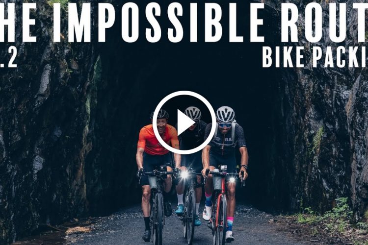Two cyclists are riding through a dark tunnel surrounded by rocky walls, with greenery visible on the sides. The text overlay reads "THE IMPOSSIBLE ROUTE EP. 2 BIKE PACKING," and a play button icon is centered on the image, indicating a video.
