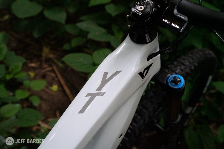 A close-up view of a white mountain bike frame featuring the logo "YT" on the top tube. The background consists of lush green foliage, highlighting the bike's design and setting. The handlebars and front fork are visible, showcasing details such as the headset and cable routing.