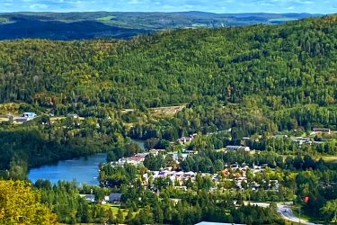 A panoramic view of a lush, green landscape featuring rolling hills and a small town nestled beside a lake. The sky is partly cloudy, and in the foreground, there is a clear view of a building and parked vehicles along a road. The scene showcases the natural beauty of the area, with a mix of trees and agricultural land. Velo Edmundston Trail mountain bike trail.