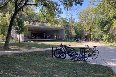 A modern concrete building with an open design situated in a lush, green park. In the foreground, several bicycles are parked on a bike rack, while people can be seen enjoying the space outside the building. The scene is framed by trees, with sunlight filtering through the leaves on a clear day. Coler Mountain Bike Preserve mountain bike trail.