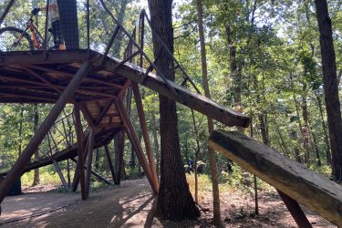 A metal structure elevated above the ground with a ramp leading down to the forest floor, surrounded by trees and greenery. A person can be seen on the platform with a bike. Coler Mountain Bike Preserve mountain bike trail.