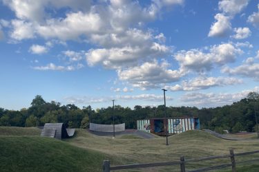 A landscape view of a park featuring a modern skateboarding area with hills and ramps. The sky is mostly sunny with scattered clouds. In the background, there is a colorful structure displaying a mural, surrounded by trees. A wooden fence is visible in the foreground. Railyard Bike Park mountain bike trail.