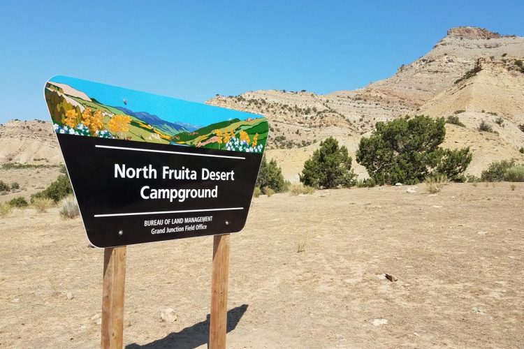 A colorful sign for the North Fruita Desert Campground, featuring artistic designs of mountains and flowers. The sign is set against a backdrop of dry, rocky terrain and hills under a clear blue sky.