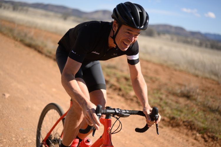 A cyclist wearing a black jersey and helmet rides a red bike along a dirt path, smiling in the sunlight. In the background, there are rolling hills and a clear blue sky, creating a vibrant outdoor scene.