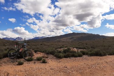 A mountain bike parked on a rocky outcrop with a landscape of shrubs and dry terrain in the foreground, set against a backdrop of distant mountains and a partly cloudy blue sky. Middelbergfarm mountain bike trail.