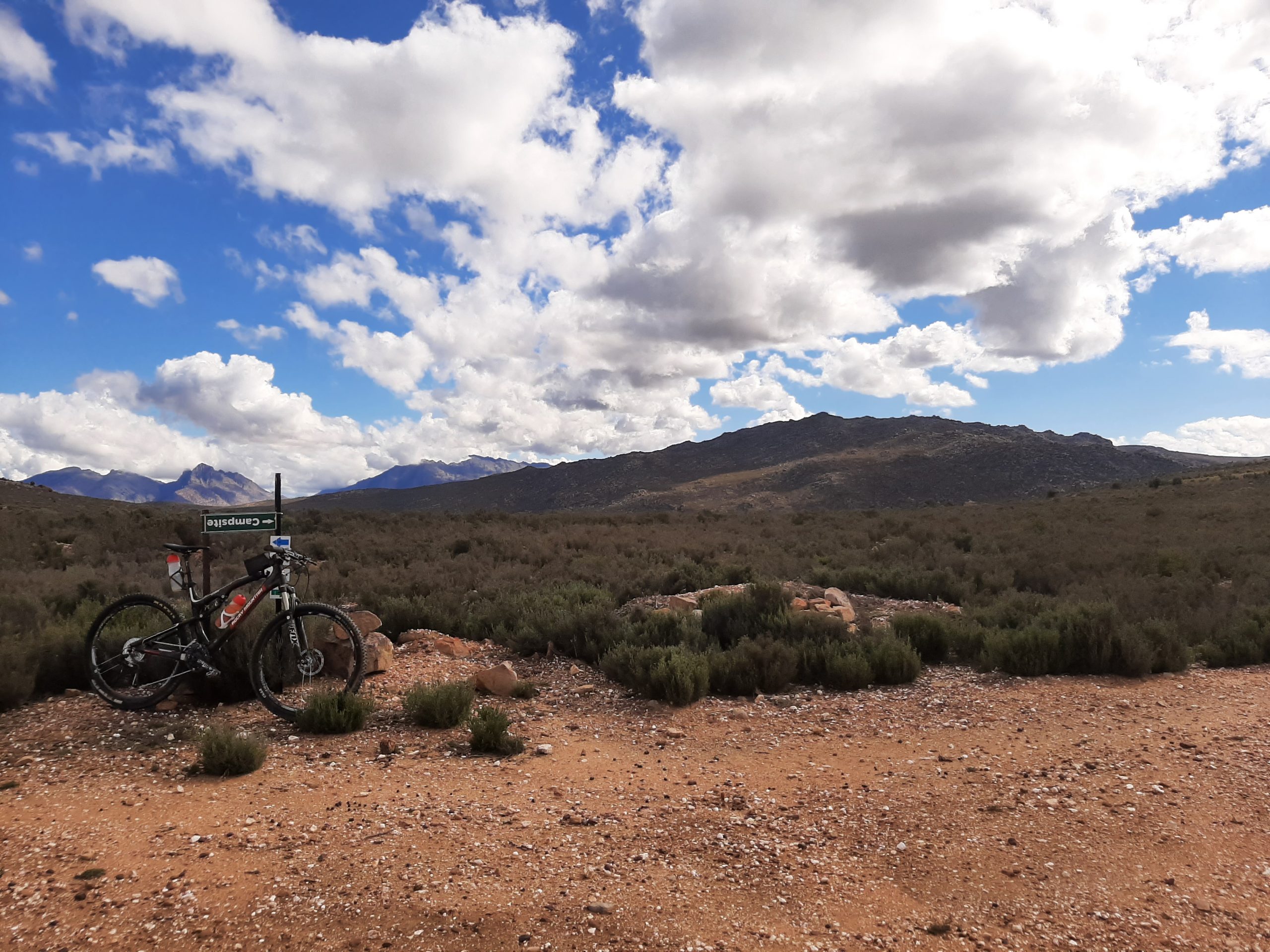 A mountain bike parked on a rocky outcrop with a landscape of shrubs and dry terrain in the foreground, set against a backdrop of distant mountains and a partly cloudy blue sky. Middelbergfarm mountain bike trail.