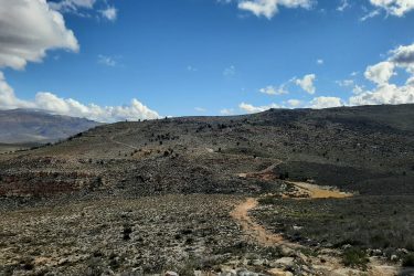 A wide landscape view of rocky terrain under a blue sky with scattered clouds. A dirt path winds through the foreground, leading towards a hilly area in the background, which is dotted with sparse vegetation and boulders. Middelbergfarm mountain bike trail.