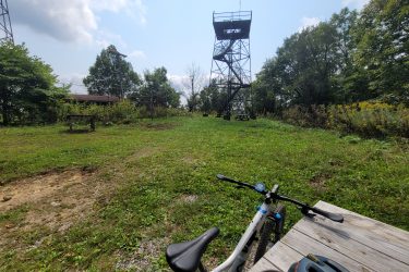 A scenic view of a grassy area with a picnic table and a mountain bike in the foreground. In the background, there is a tall observation tower surrounded by trees under a partly cloudy sky. The setting appears to be a recreational area ideal for outdoor activities. Frozen Head State Park Fire Tower Trail mountain bike trail.
