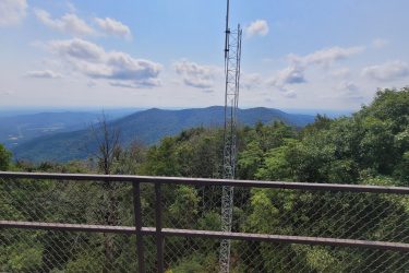 A panoramic view of rolling mountains under a clear blue sky, with scattered clouds. In the foreground, a metal railing with a fence provides a vantage point over the lush greenery and hills. A communication tower is visible to the left, blending into the natural landscape. Frozen Head State Park Fire Tower Trail mountain bike trail.