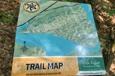 A colorful trail map displayed on a sign, featuring the layout of trails in Haw Ridge Park, Oak Ridge. The map includes various trail routes, parking areas, and notable landmarks, with a compass rose for orientation. The sign also has a warning to dial 911 for emergencies. Surrounding foliage indicates a natural, wooded environment. Haw Ridge Park mountain bike trail.