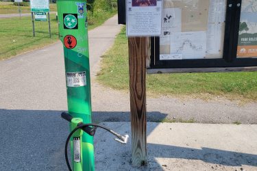 A green bike repair station with an air pump and tools stands next to a wooden information board displaying a map and trail guide for the Melton Lake Greenway. Nearby signs indicate park rules and trail information. The area is grassy with a paved path visible in the background, and power lines stretch across the horizon. Haw Ridge Park mountain bike trail.