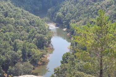 Aerial view of a winding river surrounded by lush green forests and hills, with a prominent rocky outcrop in the foreground. The scene captures the beauty of nature, highlighting the vibrant foliage and the serene waterway below. Grand Gap mountain bike trail.