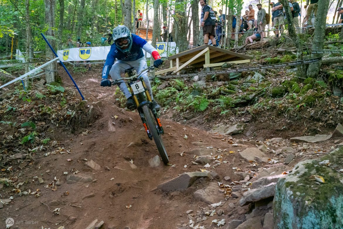 A mountain biker navigates a rocky, dirt trail in a forested area during a race, with spectators watching in the background. The biker, wearing a helmet and protective gear, leans into a turn, showcasing skill and control on the rugged terrain.