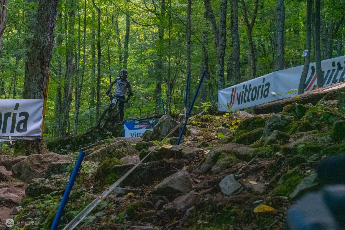 A mountain biker navigates a rocky trail in a wooded area, with banners featuring the logo "Vittoria" visible in the background. The scene captures the challenge of off-road biking, surrounded by lush green trees and an uneven terrain.