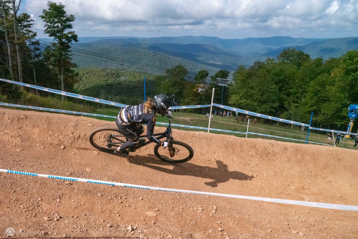 A cyclist in a black and yellow jersey navigates a dirt track with a curved berm, set against a backdrop of rolling green hills and a cloudy sky. The scene is part of a mountain biking event, with spectators visible in the distance and ski lift towers in the background.