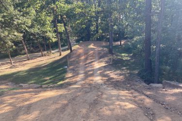 A dirt path winding through a wooded area, with trees lining either side. The sun casts dappled shadows on the ground, highlighting the trail's gradual incline. There are a couple of smaller paths intersecting in the background, creating a peaceful natural scene. Coler Mountain Bike Preserve mountain bike trail.