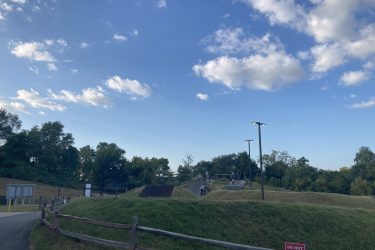 A view of a park featuring grassy hills, paved paths, and a skatepark area. The sky is partly cloudy and blue, with a few trees in the background. Signs and lighting are visible along the path, and some people can be seen enjoying the space. A "DO NOT ENTER" sign is posted in the foreground. Railyard Bike Park mountain bike trail.