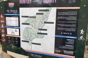 Map and information sign for the Huntley Gravity Zone at Oz Trails in Northwest Arkansas. The sign includes trail descriptions, difficulty levels, etiquette for soft surface trails, and contact information for local authorities. Features various trail names and distances. Visual elements include icons for biking and walking, along with sponsor logos. Little Sugar Creek mountain bike trail.